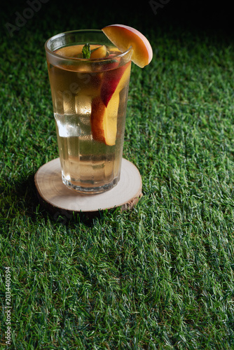 A glass of refreshing peach ice tea with mint and fruit slices placed on a rustic wood slice coaster over lush green grass