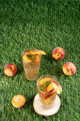 High angle view of two glasses of cold peach tea with fresh mint, scattered ripe peaches around on a green grass lawn