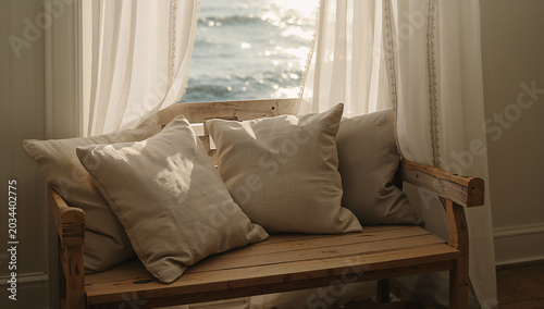 Comfortable woven bench with plush decorative pillows in front of a sheer curtained window overlooking the ocean with beach house