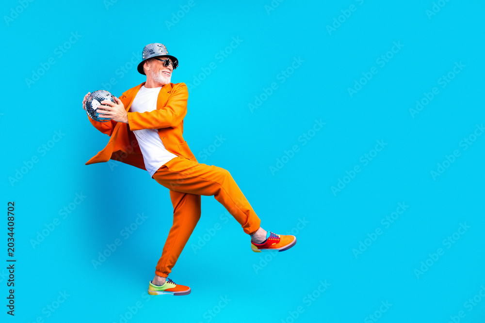 Naklejka premium Senior man in bright orange suit and hat holding a disco ball while dancing against a blue background — playful fashion, retro party and energetic movement
