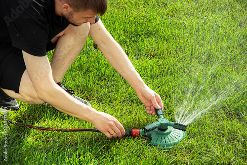 Male gardener adjusts a green lawn sprinkler on a vibrant green lawn, water spraying in arcs, showcasing a sunny outdoor setting with fresh grass
