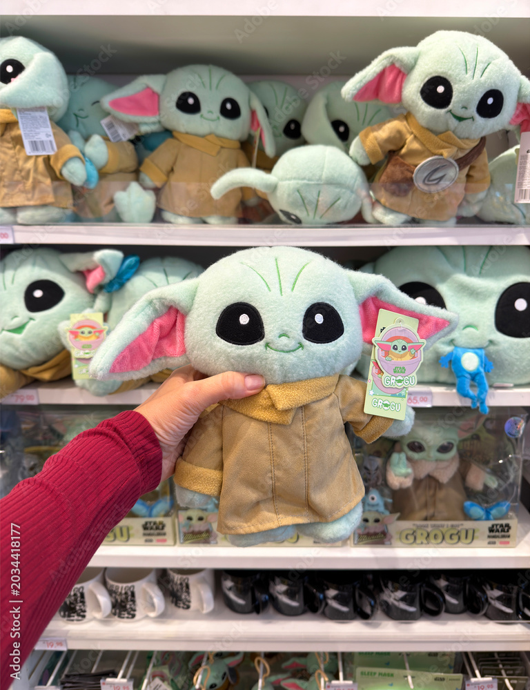 Naklejka premium Dubai, UAE - 24 may 2026: Grogu plush toy from Star Wars franchise held inside retail store with multiple character dolls on shelves, highlighting pop culture merchandise and fan shopping trend