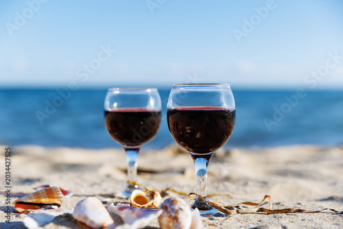 Two glasses of red wine on sandy beach with ocean horizon seashells and soft blur background summer relaxation travel lifestyle concept