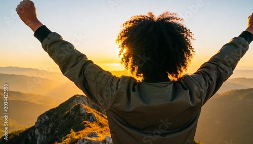 Back view of a person with curly hair raising arms in triumph on a mountain peak at sunset. Success and achievement concept. Hiker reaching the summit during golden hour. Outdoor adventure