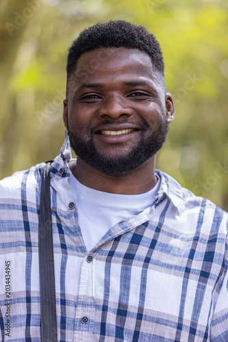 Man smiling, posing in woods wearing plaid shirt over white tee, shoulder-strap and mask-ear-loop