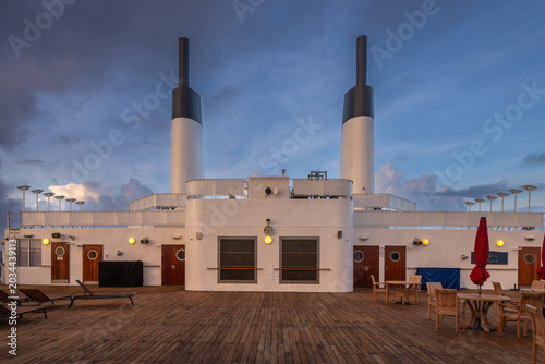 Peaceful cruise ship deck scene featuring wooden flooring, empty lounge chairs, tables with red umbrellas, and dramatic sunset clouds over the ocean. Captures a serene moment of luxury travel at sea, 