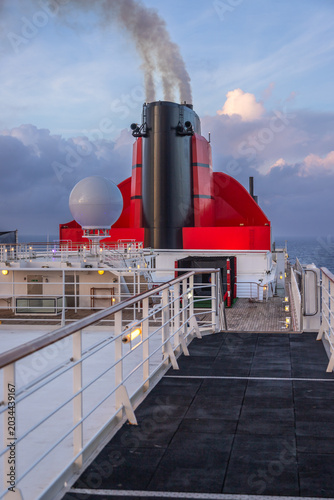 Close-up view of a large red and black cruise ship funnel releasing smoke into the sky at dusk. Captured aboard a luxury ocean liner, this maritime scene conveys the power and scale of modern cruise t