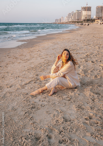 A young woman with long, thick hair sits barefoot on the sand of a city beach at sunset. In the background are the Mediterranean coastline, gentle waves, and the cityscape.