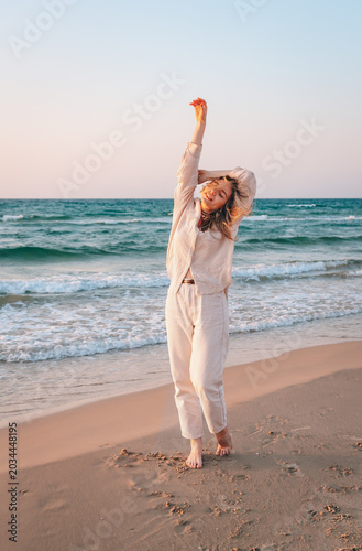 A happy young woman with long hair dances barefoot on a sandy beach at sunset. She is wearing a light, casual outfit consisting of a linen button-down shirt and matching trousers. She poses with her a