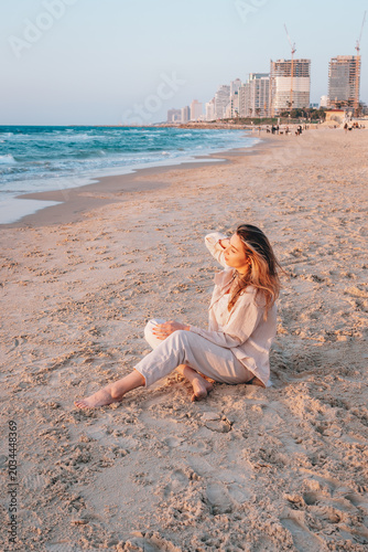 A young woman sits on a sandy beach by the sea at sunset. The warm atmosphere, light, and relaxing ambiance, along with the seascape and city coastline, create a backdrop for a tranquil summer mood.