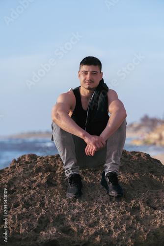 A young man of athletic build sits on a rock by the sea. Full-length portrait.