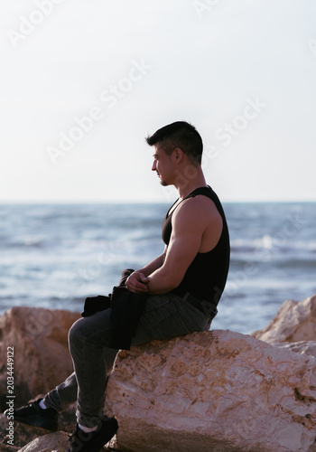 Pensive young man in black tank top sitting on rocks by the sea, profile view side shot.