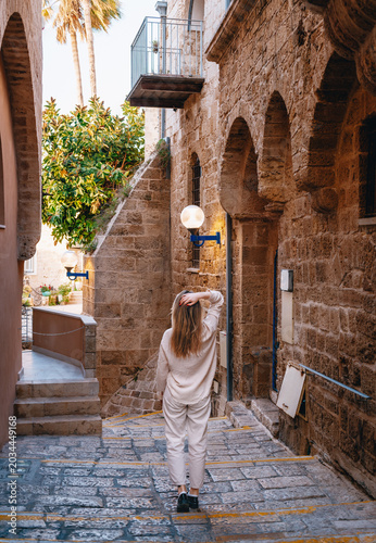 A back view of a young woman with long hair walking through a scenic narrow stone alley in the historic Old City of Jaffa, Israel.