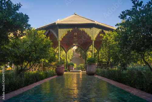 Elegant Moroccan garden pavilion in Marrakech with reflective cool mosaic path, tropical greenery and warm sunlight creating peaceful luxury atmosphere.