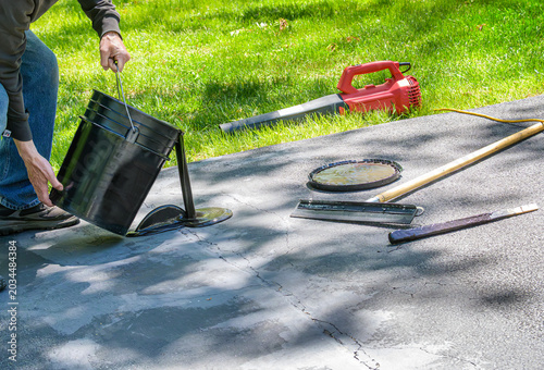 Foto Man pouring blacktop sealer on driveway cracks with spreader and blower in backg