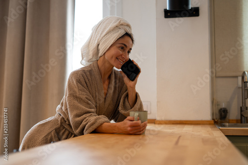 Foto Smiling woman in bathrobe with towel on head talking on smartphone while holding