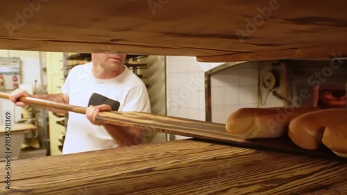 Professional baker using a long wooden peel to remove hot, freshly baked bread from a traditional oven in a commercial kitchen setting