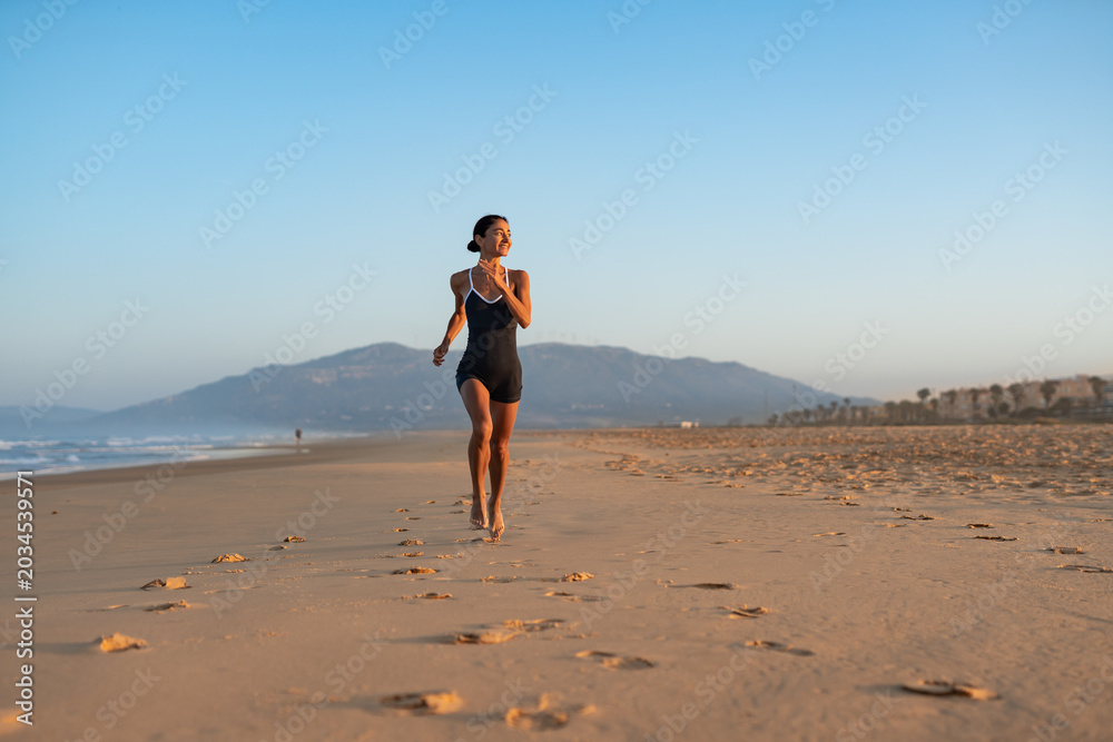 Naklejka premium Barefoot woman jogging along wide sandy beach at sunrise, mountain backdrop, long trail of footprints, healthy outdoor workout, serene coastal landscape