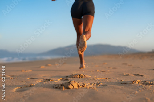 Bild auf Leinwand Back view close-up of athletic woman jogging barefoot on beach at sunrise, sand flying from her foot with every step, warm morning light and soft mountain horizon in background