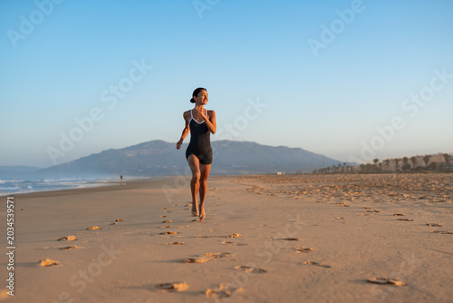 Barefoot woman jogging along wide sandy beach at sunrise, mountain backdrop, long trail of footprints, healthy outdoor workout, serene coastal landscape