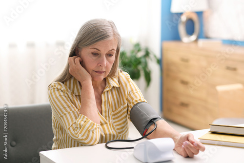 Senior woman measuring blood pressure at white table indoors
