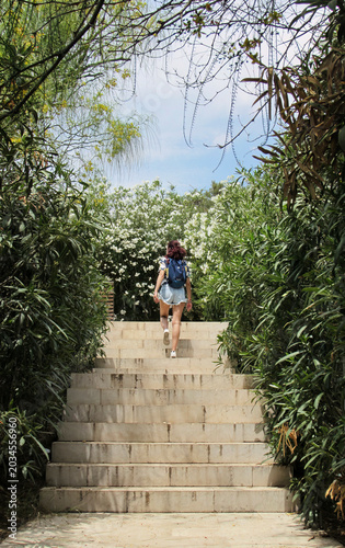 Young woman with backpack climbing up a staircase framed by dense oleander and Mediterranean trees in the gardens of Castillo de Santa Bárbara, Alicante.