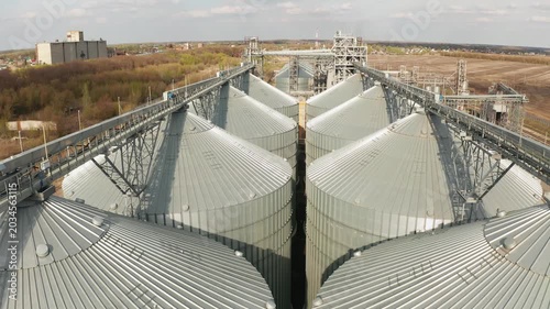 Aerial drone footage flying over rows of large steel grain silos. Industrial agricultural complex for storing and processing grain, wheat, or corn. Modern agribusiness and food production facility