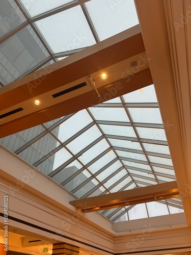 A low-angle view looking straight up at a large, pitched glass skylight ceiling inside a modern building. Large wooden or metal support beams featuring recessed warm lighting span across the glass str