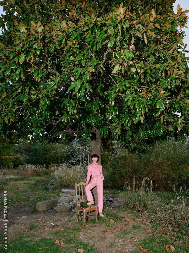 Fototapeta premium Young girl in pink overalls sits on a wooden chair beneath a lush tree in an open park, capturing a serene moment of solitude and nature, with soft light filtering through leaves.