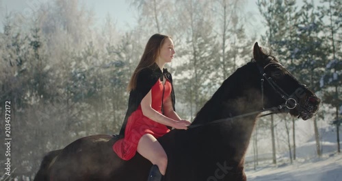 Beautiful young woman in red dress riding a black horse in winter