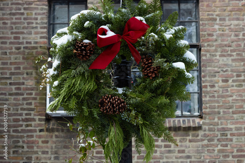 christmas wreath with pine cones and a red velvet ribbon bow outdoors with snow covering the top in front of a brick wall