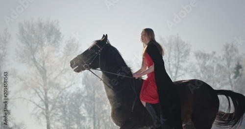 Young woman in a red dress riding a black horse in a winter forest