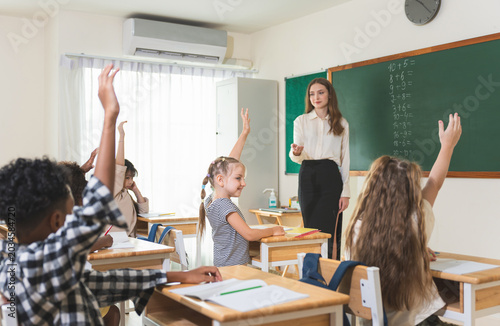 Diverse students raising hands to answer question in classroom, Teacher and elementary pupils during math lesson in school, Engaged children in classroom activity with teacher at blackboard