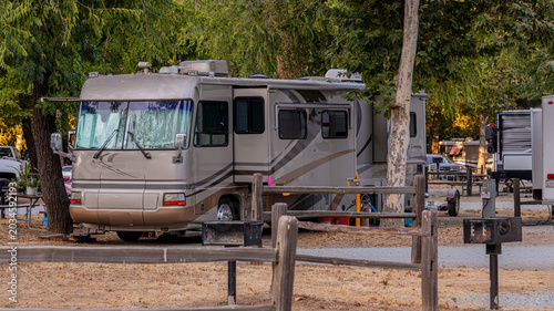 A large motorhome is parked in a serene campsite surrounded by trees. Other camping vehicles are visible in the background.