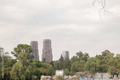 Modern city skyline with twin office towers rising above a lush urban park filled with trees and greenery under a cloudy sky