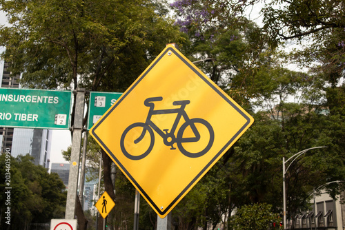 Yellow bicycle traffic sign displayed on a city street surrounded by urban greenery and modern infrastructure