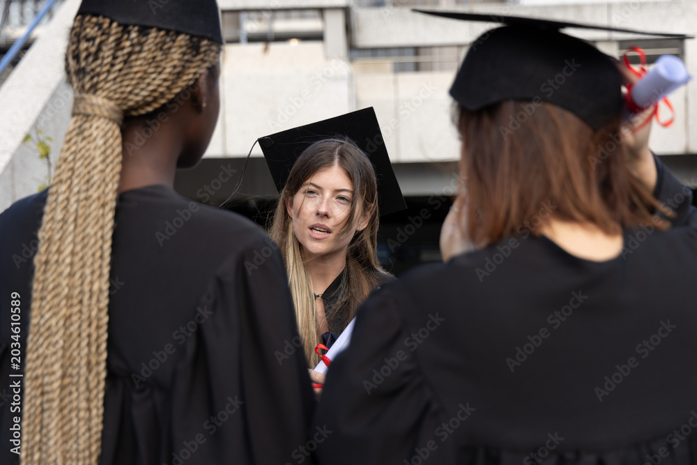Fototapeta premium Diverse women in gowns posing on campus concrete ramp, talking and holding caps, red-ribbon diploma
