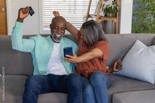 Senior couple sitting on gray sofa in living room, leaning holding smartphone and credit card