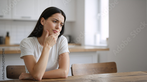 Pensive young woman sitting at a kitchen table and thinking. Worried female with hand on chin looking away in thought. Mental health and stress concept