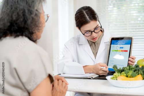 Asian woman nutritionist showing AI BMI calculator on tablet and explaining to senior patient, doctor using digital health technology for medical diagnosis, wellness planning and weight control.