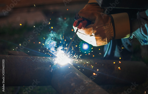 Closeup of skilled welder joining steel pipe with electric arc in dark workshop environment. Bright sparks and glowing metal create dramatic industrial scene focused on precision craftsmanship.