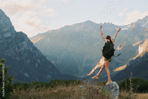Fotografija Young woman with backpack stands on a rocky ledge, arms raised in celebration, s