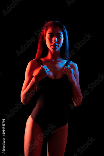 Fashion portrait striking image of a woman in a dark bathing suit, lighting color half lit in red and half in blue studio shot on black background.