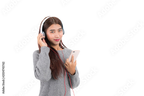 Beautiful young Asian woman is enjoying music on her mobile phone with a pair of headphone. She is standing and relaxing, wearing a gray knit sweater at studio shot isolated on white background.