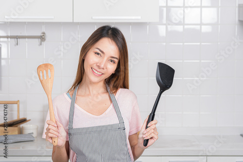 Beautiful Asian young woman joyfully poses in her white kitchen, holding cooking tool is a spatula and wooden spoon representing a love for homemade meal.