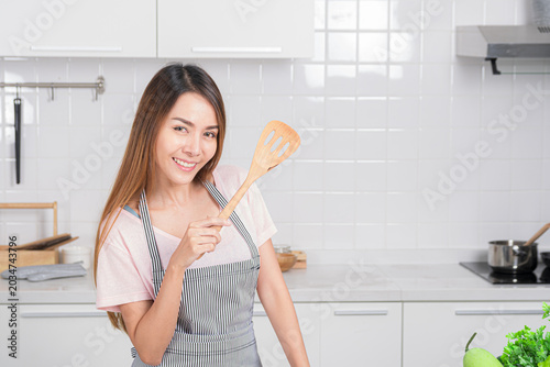 Beautiful Asian young woman joyfully poses in her white kitchen, holding cooking tool is wood spatula representing love for homemade meal.