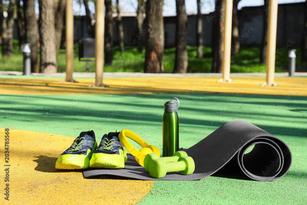 Naklejka premium Fitness mat with sneakers, headphones, water bottle and dumbbells on sports ground