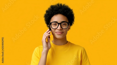 Smiling young person with curly hair and glasses having phone conversation on vibrant yellow background