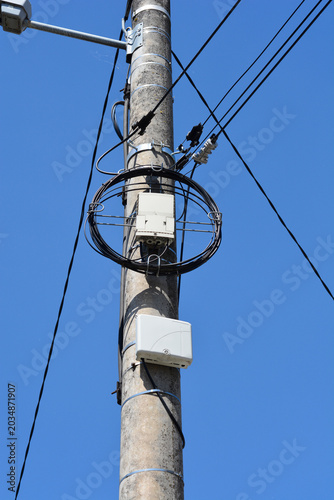 A concrete utility pole features mounted white telecommunication boxes and coiled fiber optic cables against a clear blue sky