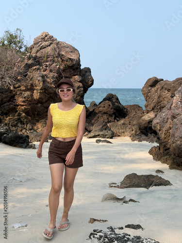 Happy Asian Thai woman enjoying a tropical beach walk, smiling amidst rugged rock formations, blue ocean, and soft sand under a clear sky.